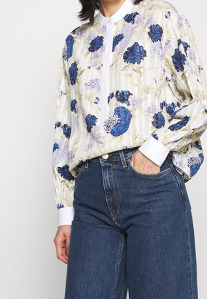 Blusa de manga larga con estampado floral en azul y púrpura, cuello y puños blancos, metida dentro de unos vaqueros de tiro alto en azul oscuro, con la mano en el bolsillo.