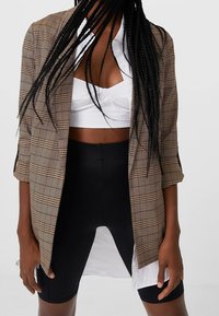 Hnědý kostkovaný blazer s přiléhavým střihem, kombinovaný s bílým zkráceným topem a černými šortkami s vysokým pasem, ukazující vrstvený vzhled.