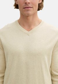 Beiger V-Ausschnitt-Pullover aus weichem, strukturiertem Strickstoff. Verfügt über enge Rippdetails am Kragen und ein geprägtes Logo auf der Brust.