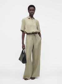 Personne portant une chemise beige ample boutonnée et un pantalon à jambes larges, tenant un sac à main noir, debout devant un fond clair uni.