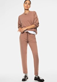 Ensemble de sweat-shirt et pantalon marron en tissu doux. La tenue présente une coupe décontractée, des manches resserrées et une taille avec cordon de serrage. Des chaussures noires complètent le look.