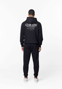 Man die met zijn rug naar de camera staat, draagt een zwarte hoodie en broek met witte tekst "CLSR LDN" op de achterkant, en witte sneakers, tegen een effen witte achtergrond.