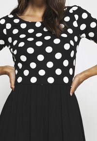 Musta mekko valkoisilla pilkuilla, pyöreällä pääntiellä ja lyhyillä hihoilla. Valuva hame, jossa vyötäröllä kerättyjä yksityiskohtia.