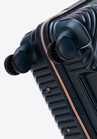 Gros plan du coin d'une valise rigide noire avec des roues doubles pivotantes et une fermeture éclair couleur or rose sur fond blanc.