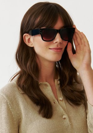 Melni pārmērīgi lieli saulesbrilles ar tumšu gradientu lēcām, kvadrātveida rāmi un biezām kājiņām, ko valkā persona ar gariem brūniem matiem un bēšu augšdaļu.