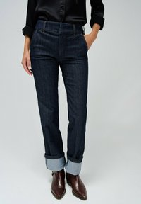 Pantalones de mezclilla oscuros con una cintura alta, que presentan una pierna estrecha y dobladillos enrollados. Combinados con botas de tobillo marrones.