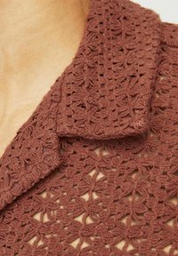 Blusa marrón de ganchillo con cuello, que presenta un patrón floral. La textura es de punto abierto y ligera, mostrando intrincados detalles.