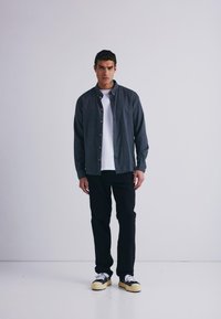 Camisa de botón gris oscuro sobre una camiseta blanca, combinada con pantalones negros y zapatillas en blanco y negro. Tejido suave con un corte relajado.