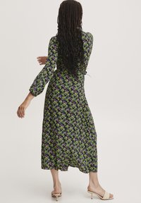 Lungo vestito floreale con uno sfondo nero caratterizzato da piccoli fiori viola e verdi, maniche a sbuffo e una silhouette fluida. Indossato con tacchi leggeri.