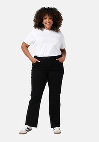 Pantalons noirs taille haute avec poches avant, assortis d'un t-shirt blanc portant de petits motifs en forme de cœur. Les chaussures sont des baskets blanches avec des accents noirs.
