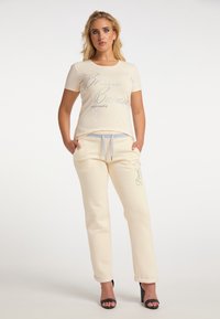 Hellbeige T-Shirt mit silberner Schrift, kombiniert mit passenden Jogginghosen, die einen Kordelzug und ein Logo-Detail am Bein aufweisen. Glatte Stofftextur.