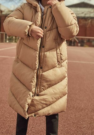 Manteau matelassé beige avec une fermeture éclair à l'avant, un matelassage en chevrons et une capuche. Le manteau possède des poignets élastiques et des poches latérales, porté avec un pantalon foncé.