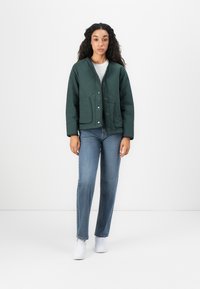 Veste matelassée verte forêt avec un col rond, des boutons-pression et deux poches avant, portée avec un jean bleu clair et des baskets blanches.