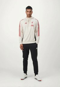 Hvid atletisk pullover med røde accenter, med hætte og lynlås, parret med sorte bukser og hvide sneakers. Adidas-branding synlig.