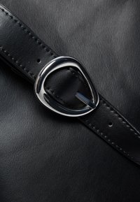 Schwarzes Lederarmband mit metallic-silberner Schnalle, versehen mit sichtbaren Nähten und einer strukturierten Oberfläche. Die Schnalle hat eine Tropfenform.