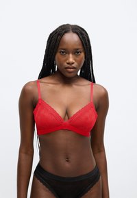 Bralette vermelho de renda com decote em V, alças ajustáveis e padrão com logótipo. Cuecas pretas de renda com bordas recortadas complementam o conjunto.