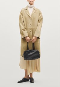 Tan suède lange jas met een oversized pasvorm, zwarte geweven handtas, crèmekleurige coltrui, geplooide beige rok en zwarte muiltjes met spitsen.