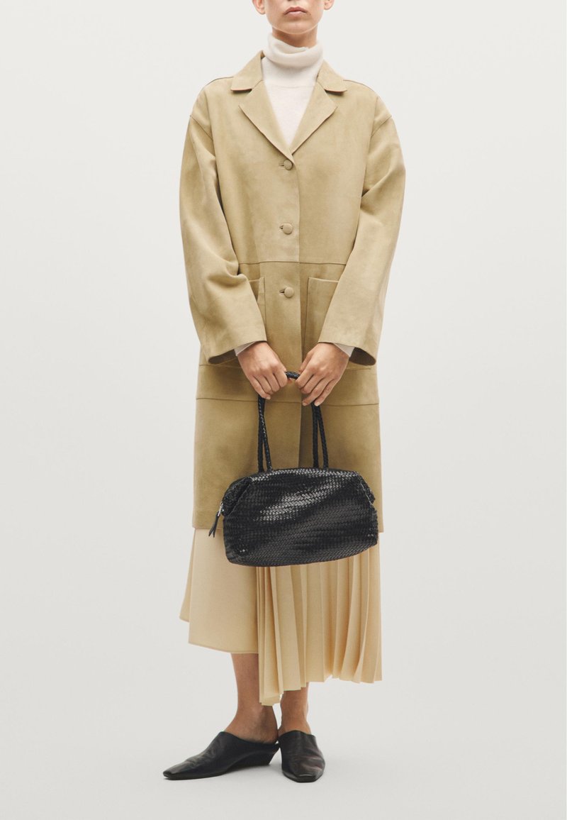 Manteau long en suede camel avec coupe oversized, sac à main noir tissé, col roulé crème, jupe beige plissée et mules noires à bouts pointus.