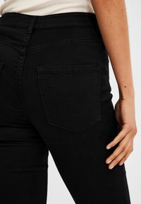 Mustat farkut, joissa on lähikuva takataskusta ja vyötäröstä. Kangas on sileä ja siinä on hienovaraisia tikkaus yksityiskohtia.