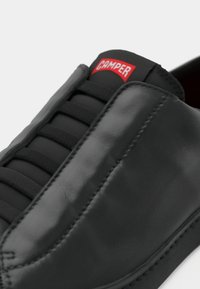Schwarzer Ledersneaker mit elastischen Schnürsenkeln, auffälligem roten "Camper"-Logo, glatter Textur, rund zulaufender Spitze und sichtbaren Nahtdetails.