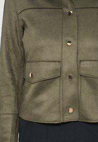 Χακί πράσινο σακάκι με τέσσερα γυαλιστερά χρυσά κουμπιά, δύο μπροστινές τσέπες με καπάκι και μακριά μανίκια, φαίνεται από το στήθος έως τη μέση.