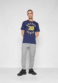 Námořnické tričko s žlutým potiskem "GOLDEN STATE 30", v kombinaci se šedými tepláky a černými sportovními botami, postavené na jednobarevném pozadí.