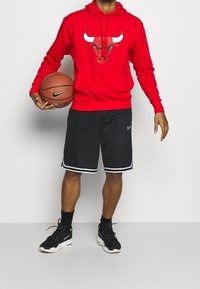 Červená mikina s kapucí s grafikou býka, černé basketbalové kraťasy s bílými pruhy a černé tenisky se zelenými akcenty. Drží basketbal.