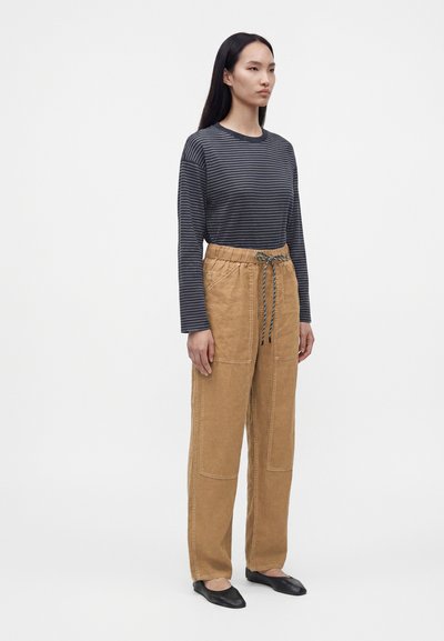 Femme debout portant une chemise à manches longues rayée bleu marine, un pantalon camel taille haute avec un cordon de serrage, et des chaussures plates noires sur fond blanc.