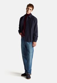 Camisa azul marino de botones con cuello, combinada con un suéter de cuello alto burdeos y jeans holgados de color azul claro. Calzado sencillo en color oscuro.