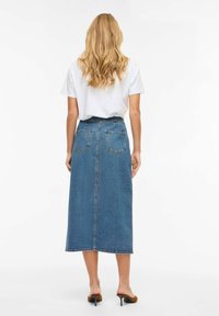 Maxirock aus Denim, mittleres Blau, hochtailliert mit zwei Gesäßtaschen, gerader Schnitt, roher Saum. Modell trägt ein weißes T-Shirt und braune offene Sandaletten.