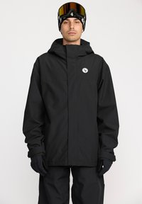 Veste imperméable noire avec capuche, fermeture éclair devant, et grand écusson logo. Le modèle porte des gants noirs et des lunettes de ski, posé devant un fond neutre.