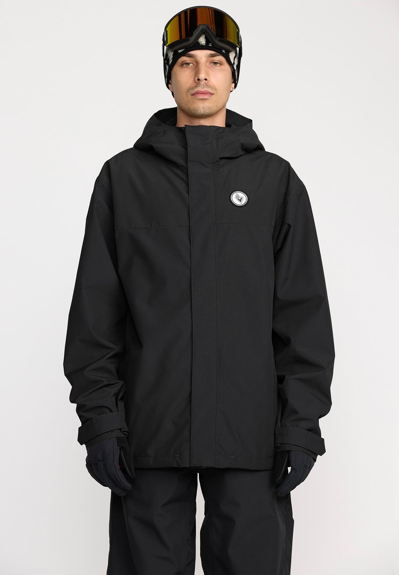 Veste imperméable noire avec capuche, fermeture éclair devant, et grand écusson logo. Le modèle porte des gants noirs et des lunettes de ski, posé devant un fond neutre.