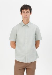 Camisa de manga corta verde claro con cuello abotonado, textura sutil y dobladillo curvado. Lleva puestos pantalones marrones.