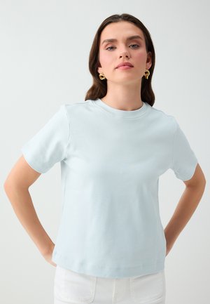 Vrouw met bruin haar draagt een lichtblauw T-shirt met korte mouwen en een witte broek, poseert met handen in de zij tegen een effen lichte achtergrond.