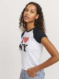Īsām piedurknei, saīsinātā T-kreklā ar baltu ķermeni un melniem raganļu āmuriem, uz kura ir neuzmanīgi sarkani un melni burti "I ♥ NY".