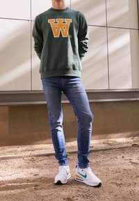 Zelená mikina s žlutým logem 'W', modré džíny a bílé tenisky s akcenty v tealové a žluté barvě, stojící na písečné ploše.