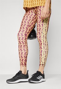 Leggings s ponavljajućim uzorkom perja u tamnocrvenoj i žutoj boji, s zelenim bočnim trakama, upaireni s crnim sportskim cipelama.