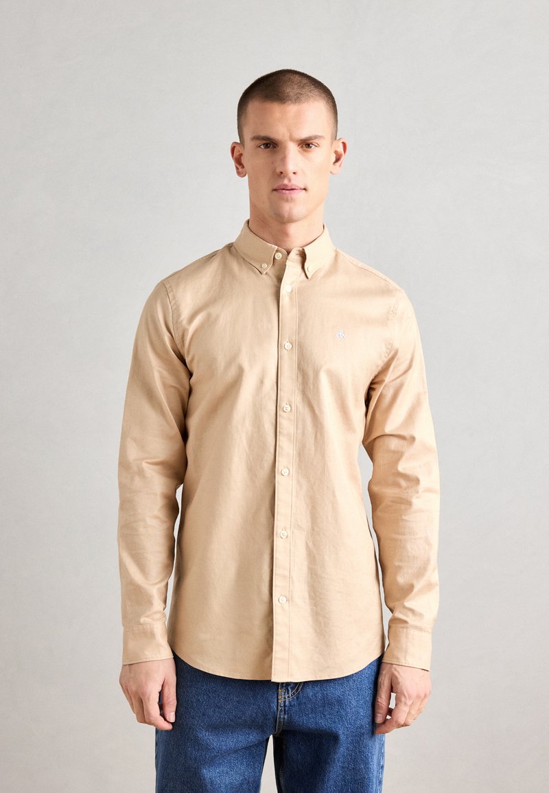 Chemise beige à manches longues avec un col boutonné, en tissu lisse, arborant un petit logo sur la poitrine, accompagnée d'un jean bleu.