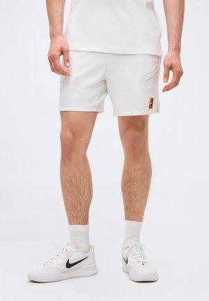 Man draagt witte sportshorts met een klein kleurrijk logo vlak bij de zoom, witte sokken en witte Nike-sneakers met zwarte swooshes.