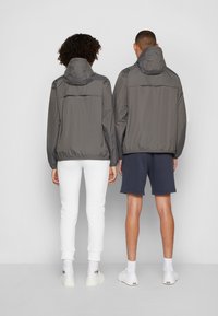 Deux personnes portent des vestes imperméables grises avec des capuches, dotées d'ourlets élastiques. L'une porte un pantalon blanc, tandis que l'autre porte un short bleu marine.