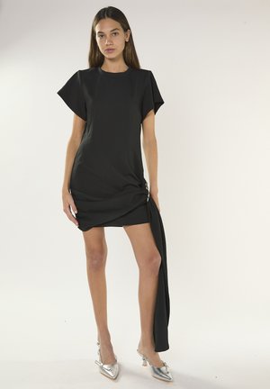 Mujer vistiendo un mini vestido negro de mangas cortas con un detalle drapeado asimétrico y zapatos de tacón alto plateados con punta, posando sobre un fondo blanco.