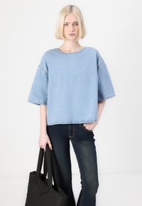 Camicetta in denim azzurro chiaro con maniche corte, scollo rotondo, bordi grezzi e silhouette quadrata, abbinata a jeans blu scuro e una borsa tote nera.