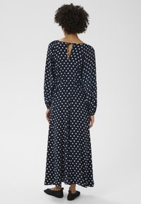 Robe bleu marine à pois avec manches longues, ouverture au dos en forme d'œillet, et silhouette fluide, associée à des chaussures noires à enfiler.