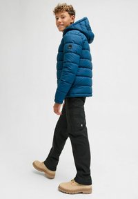 Un adolescent marche, portant une veste en duvet bleue avec une capuche, un pantalon cargo noir et des bottes beiges sur un fond clair uni.