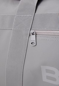 Sac de voyage gris en tissu durable, avec une fermeture éclair visible portant une étiquette "BORG" et une sangle en toile robuste. Design minimaliste avec un branding subtil.
