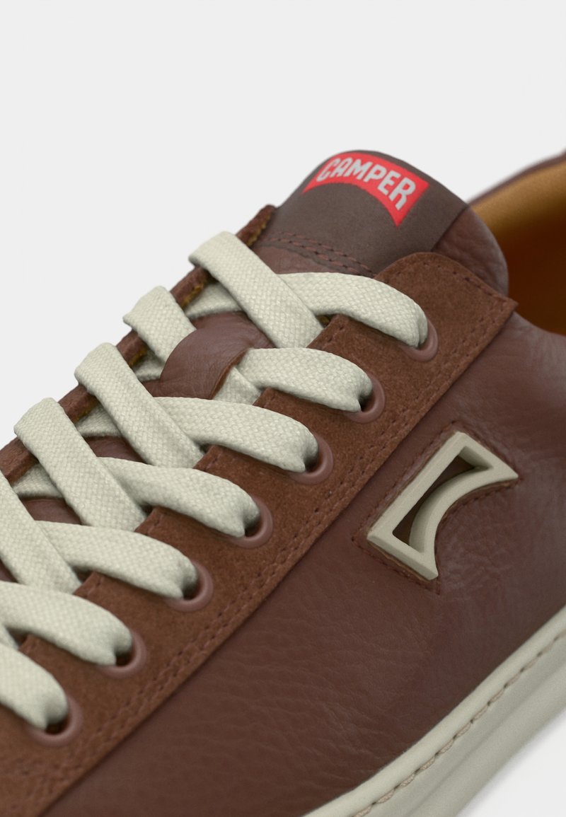Brown leather sneaker with beige laces, featuring a gray logo accent and a padded tongue. Smooth texture and classic low-profile shape.