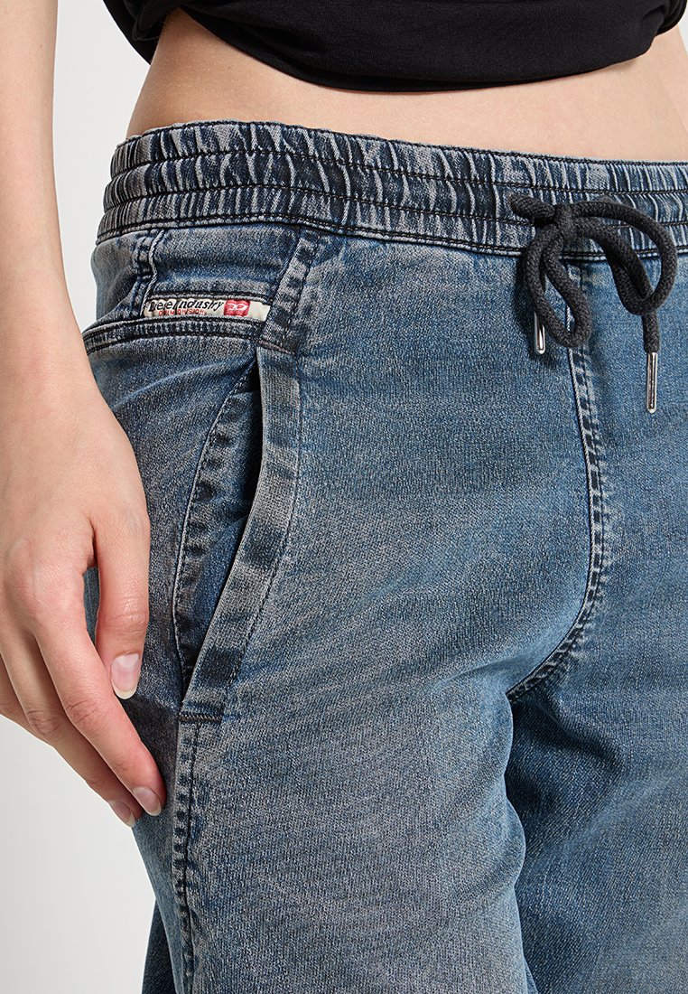 Nahaufnahme einer Person, die blaue Denim-Jogginghosen mit elastischem Bund und schwarzem Kordelzug trägt, mit einer Hand, die nahe der Seitentasche ruht.