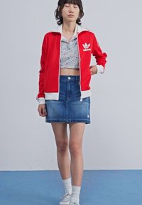 Crvena Adidas jakna s bijelim ovratnikom, cvjetni crop top i plava traper suknja. Par bijelih tenisica i čarape do gležnjeva upotpunjuju odjevnu kombinaciju.