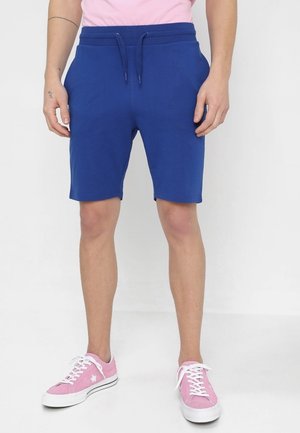 Mann trägt blaue Shorts mit Kordelzug, pinke Sneakers mit weißen Schnürsenkeln und Stern-Detail, steht vor weißem Hintergrund.