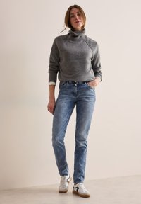 Un pull à col roulé gris avec une finition texturée, associé à un jean en denim bleu clair. Des baskets blanches avec des accents noirs complètent la tenue.